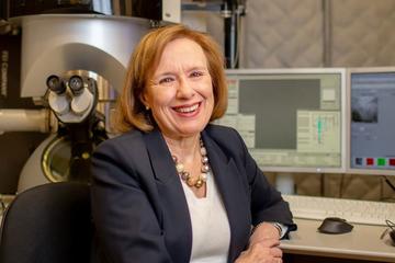 Professor Burke smiling and sitting at her lab desk with a microscope behind.
