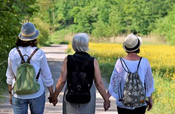 The backs of three women of mature years holding hands and walking with backpacks to an attractive rural location